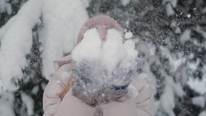 年轻女子对着镜头吹雪花。微笑的漂亮女孩，穿着白色的冬季夹克，戴着蓝色的针织帽，吹着手掌上的雪。快乐的