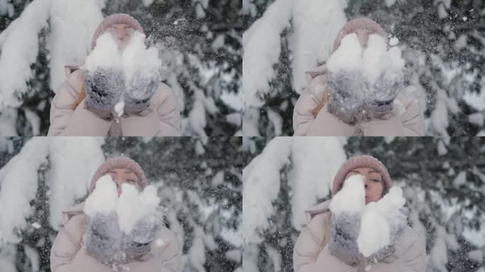 年轻女子对着镜头吹雪花。微笑的漂亮女孩，穿着白色的冬季夹克，戴着蓝色的针织帽，吹着手掌上的雪。快乐的