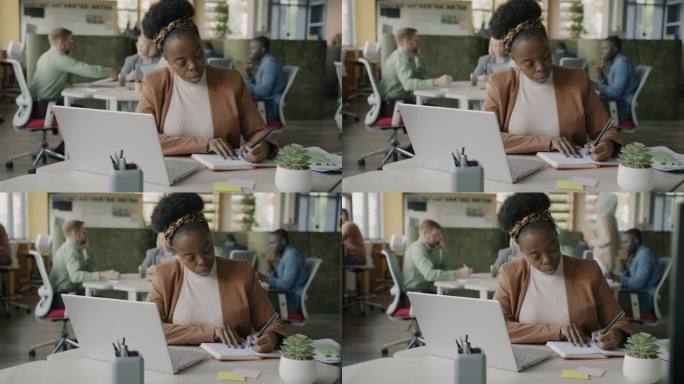 严肃的非裔美国妇女在办公室的桌子上写着笔记本电脑，看着笔记本电脑屏幕