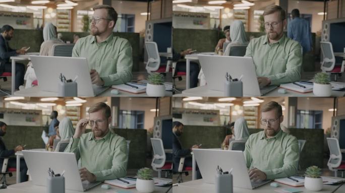 一个自信的年轻人坐在联合办公中心的办公桌前，用笔记本电脑打字，然后戴上眼镜