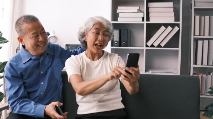 老年夫妇坐在沙发上用智能手机与家人进行视频会议和问候，花时间在家客厅一起做一些放松生活的活动。