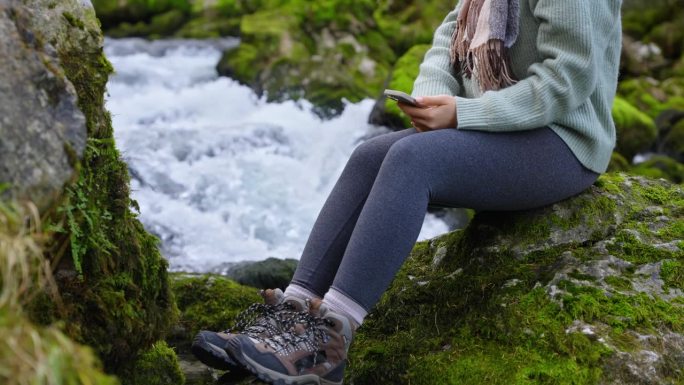 一名女子坐在森林中流经苔藓岩石的河流旁使用手机的低剖面