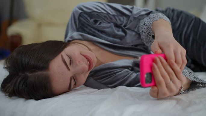特写镜头。一个穿着睡衣的女孩斜躺在卧室的床上，笑着，手里拿着手机写着短信。女孩放下手机，准备睡个午觉