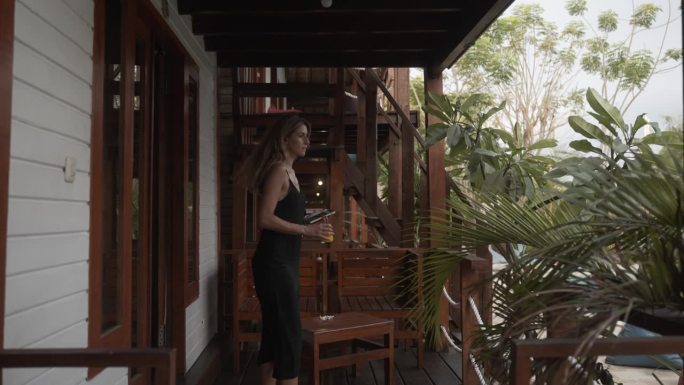 穿着黑色连衣裙的女人在阳台上放松，俯瞰着游泳池和丛林