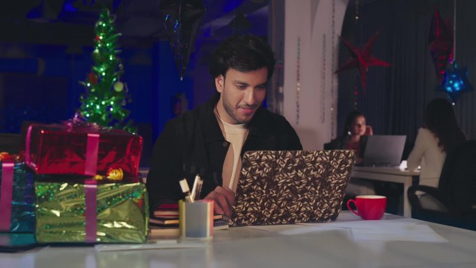 印度微笑的年轻胡须男子坐在椅子上使用笔记本电脑享受圣诞之夜庆祝搞笑派对室内办公室。快乐的帅哥在电脑上