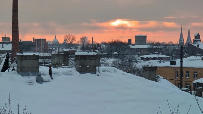 在城市工业区的屋顶上，雪花缓缓落下，太阳在灰色的冬日天空中发出橙色的光芒