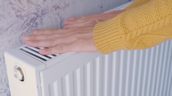 在寒冷的冬天，一名男子穿着黄色毛衣在家里的暖气上暖手。男性在暖气片上暖胳膊。供暖季节或寒冷天气的概念