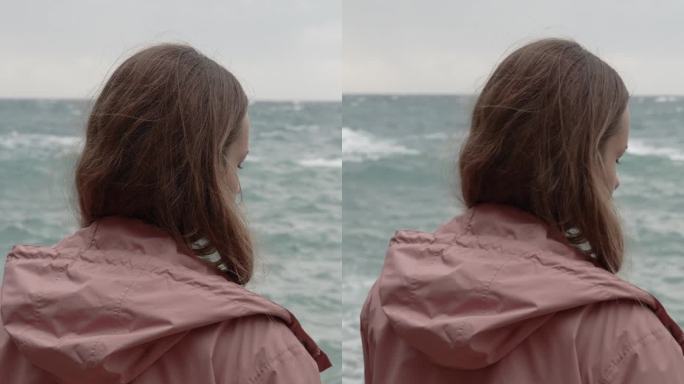 垂直视频。从背后看，一个年轻女子独自站在岩石上，面对汹涌的大海。