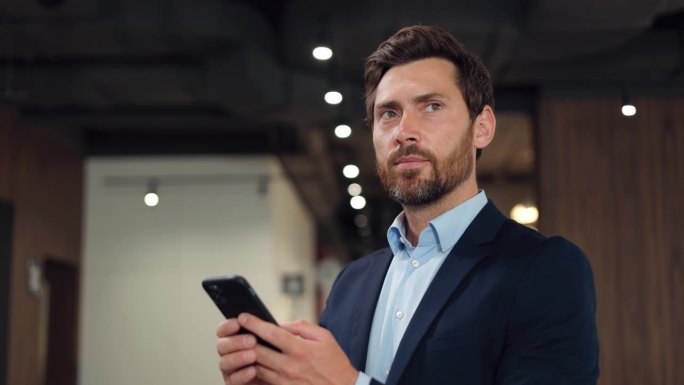 一个穿着正装、蓄着胡子的英俊白人男子站在联合办公空间里，一边用手机聊天，一边扭过头去看。黑发男士使用