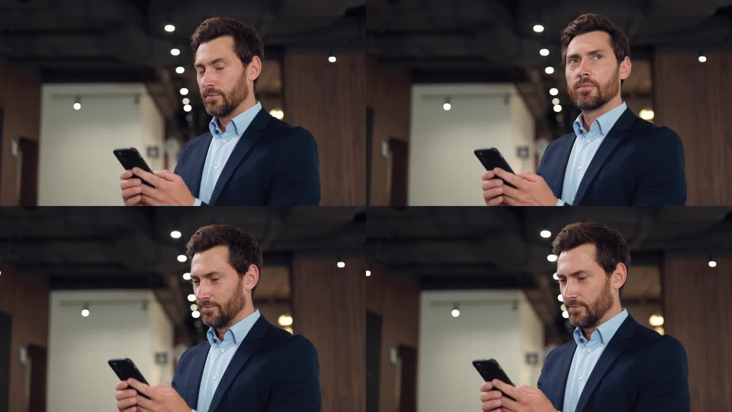 一个穿着正装、蓄着胡子的英俊白人男子站在联合办公空间里，一边用手机聊天，一边扭过头去看。黑发男士使用