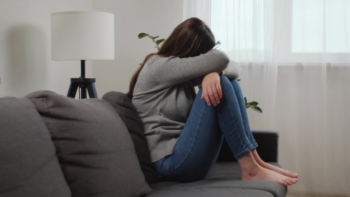 不快乐的焦虑年轻的白人女子用膝盖遮住脸坐在家里客厅的沙发上，伤心欲裂的女性在抑郁中感到绝望和焦虑，有