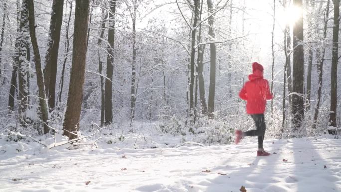 在阳光明媚、霜冻的日子里，穿着鲜红色防风夹克的越野跑女子在风景如画的雪林小路上慢跑。运动健步者和冬训