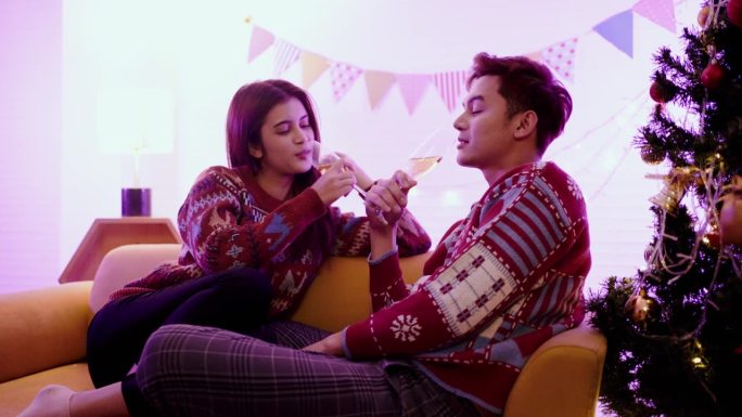 幸福的亚洲夫妇一起举杯庆祝香槟，晚上坐在沙发上，背景是圣诞树和灯光。圣诞夜的男女朋友