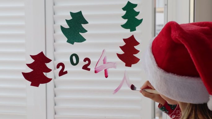 小女孩戴着红色圣诞老人帽子装饰冬季度假屋新年晚会手工纸花环和画数字2024圣诞树。儿童手工DIY家居