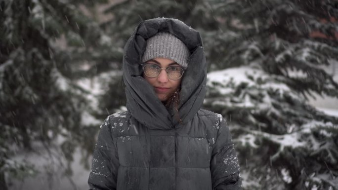 冬天下大雪，一位年轻女子站在冷杉树的背景下。一个女孩戴着眼镜，穿着带兜帽的羽绒服，站在那里看着镜头。