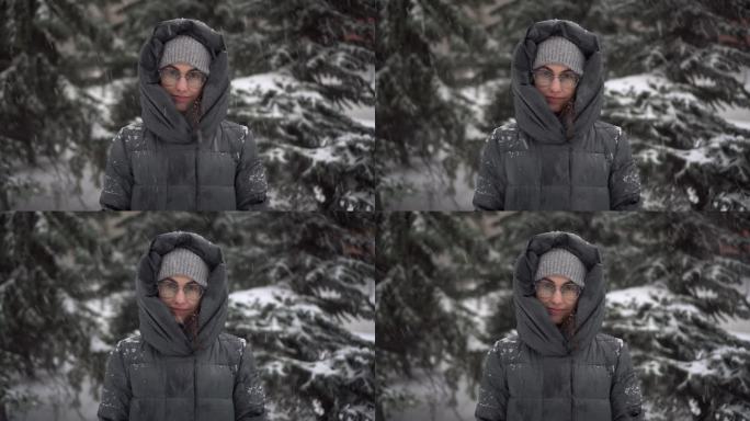 冬天下大雪，一位年轻女子站在冷杉树的背景下。一个女孩戴着眼镜，穿着带兜帽的羽绒服，站在那里看着镜头。