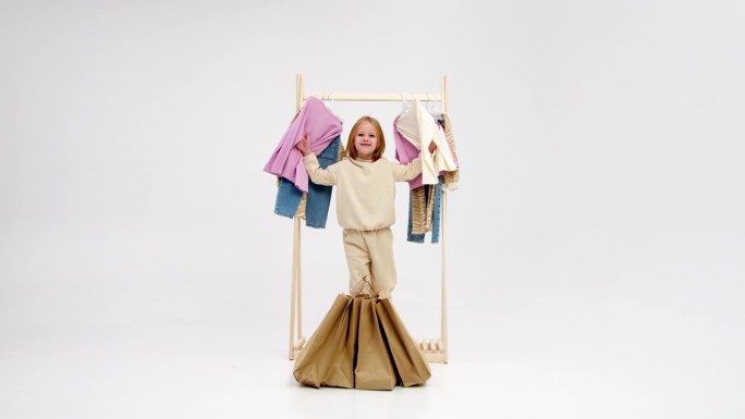 小时尚达人带着浅色背景的包裹。可爱的微笑的小女孩在衣架的背景袋享受购物。童装风格广告理念。