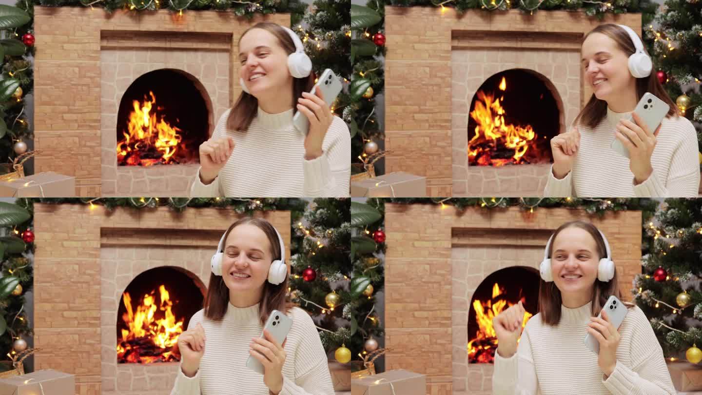 在冬天的晚上，戴着耳机的迷人女人坐在壁炉和圣诞树旁，在音乐中快乐，随着音乐的节奏跳舞。