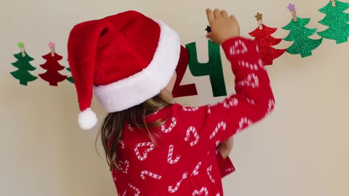 有趣，兴奋，快乐的学步女孩穿着圣诞服装，红色圣诞老人帽装饰冬季度假屋，手工纸花环数字2024。孩子们