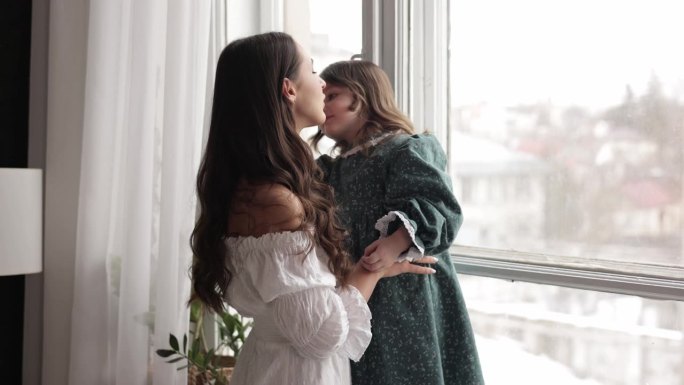 母亲的一天。年轻漂亮的妈妈和可爱的女儿在家里的全景窗边互相拥抱，深情地亲吻。快乐母性，人生价值，亲情