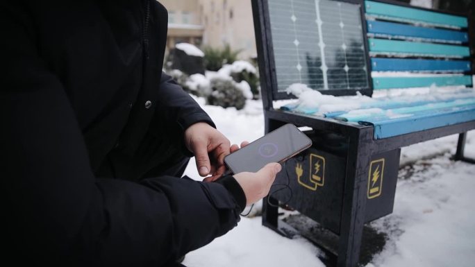 冬天，男子在户外用USB给手机充电。街道上安装太阳能电池板的公共充电长椅。替代电源。近距离