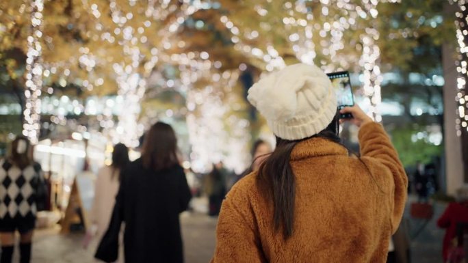 一名女游客在寒冷的圣诞夜用手机拍摄城市街道的装饰和灯光。