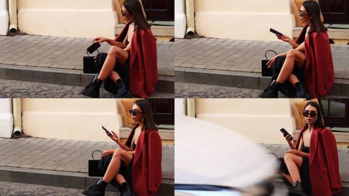 商务女孩坐在人行道上，从钱包里掏出手机。前景中有一辆汽车开过。