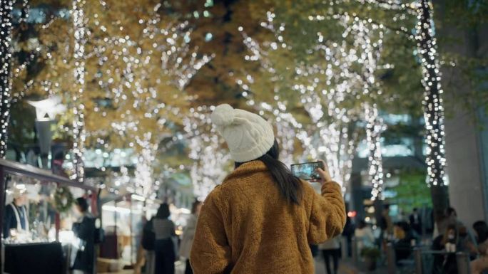 一个女人在美丽的圣诞彩灯下用手机拍照的后视图。