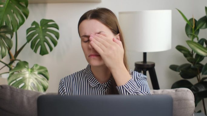 疲倦悲伤的女揉干易激的眼睛感觉眼睛疲劳紧张偏头痛后的电脑工作，疲惫的年轻女职工缓解头痛，差弱模糊的视