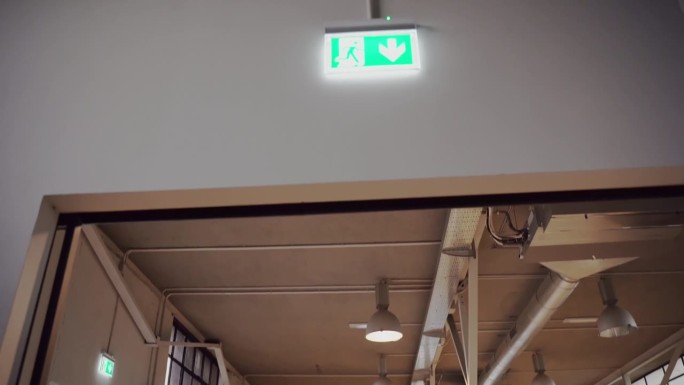 门口入口处上方悬挂绿色霓虹灯紧急出口标志，标志上发光向下的箭头和奔跑的人，指示紧急情况下的逃生方向