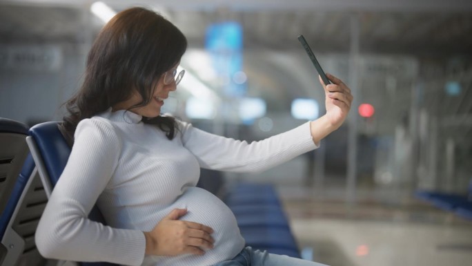 一名怀孕的亚洲女性旅客在机场候机时用智能手机与朋友视频聊天。