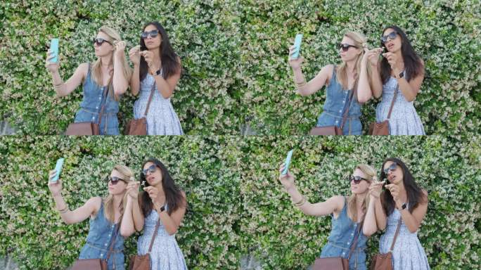 最好的朋友在度假时用智能手机自拍，头上戴着花。女孩们站在花墙前用手机分享生活照片，发短信享受夏天