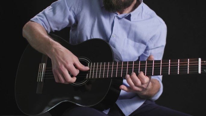 一位男音乐家手拿吉他，坐在黑暗的背景上，用手指拨动琴弦，演奏着旋律