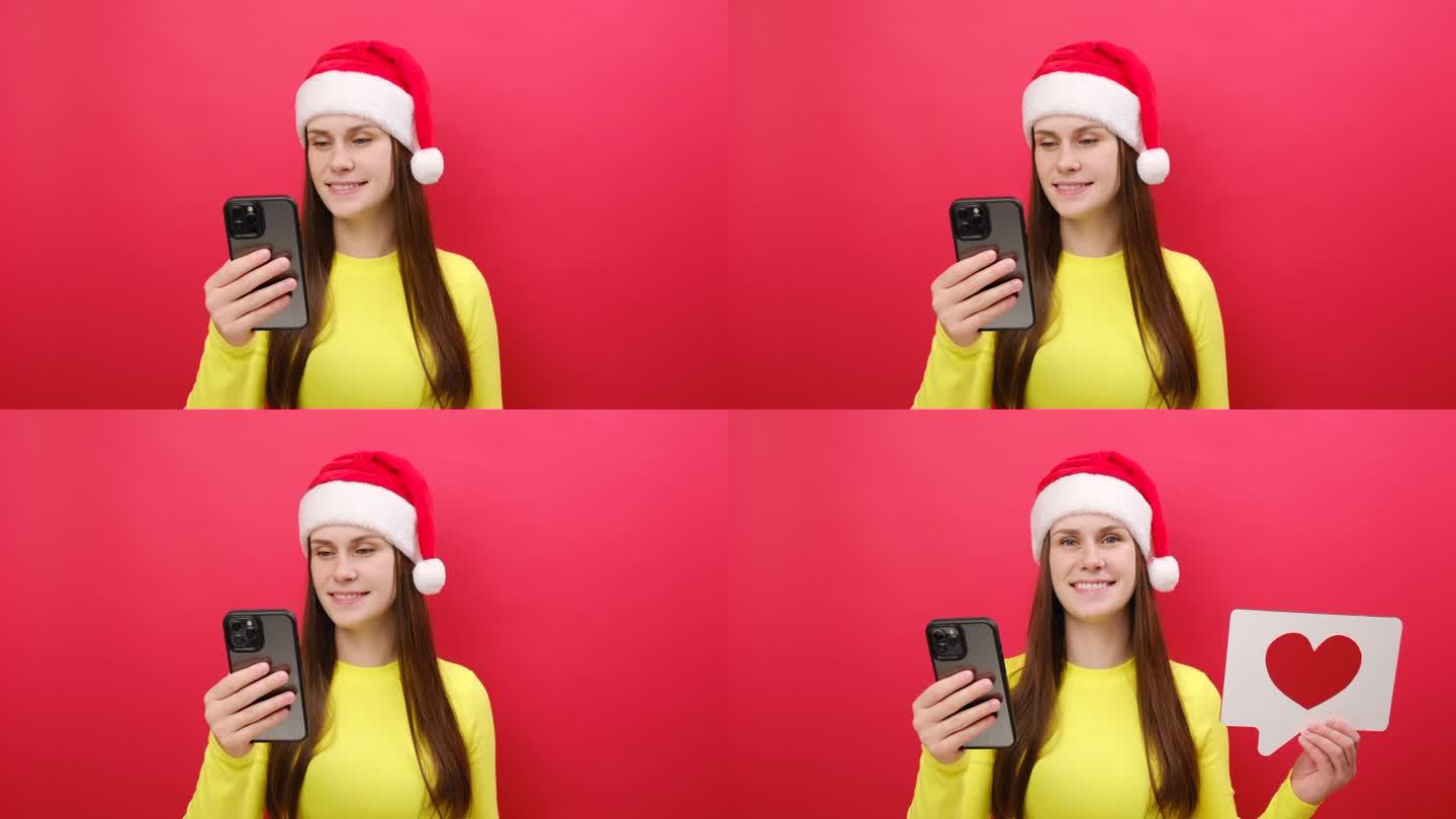 快乐的年轻女子穿着黄色的毛衣，手里拿着圣诞老人的帽子，用手机心形像图标一样的标志，在柔和的红色背景上