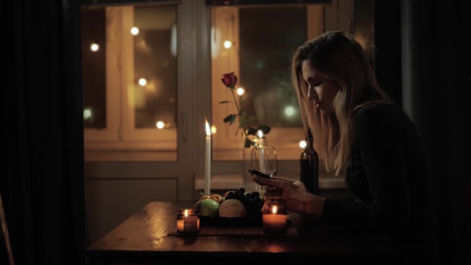 孤独的女人，失败的约会。迷人的悲伤的女人独自坐在桌子上，在美丽浪漫的环境中，点着蜡烛，喝着酒，有时看