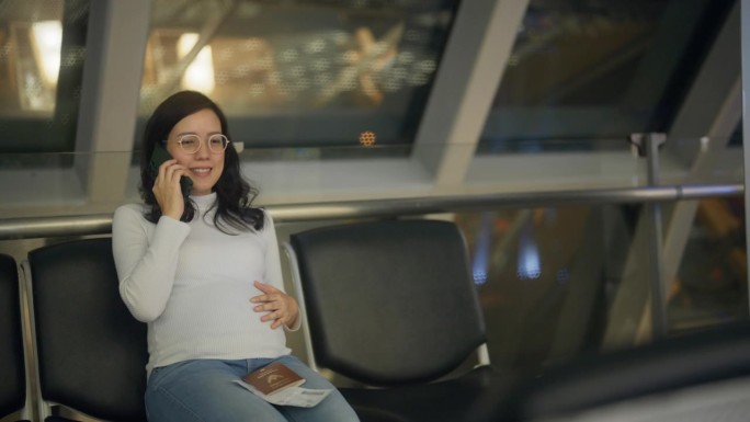 愉快的孕妇坐在沙发上等待，与家人在机场打电话