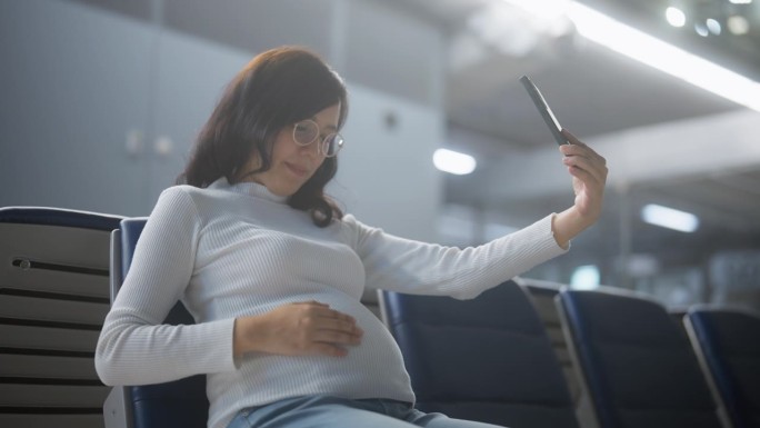快乐的亚洲孕妇在机场等候飞往国外时，用电话和视频与亲戚聊天