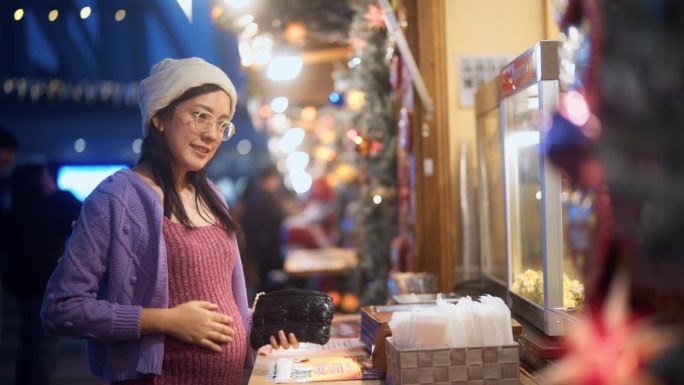 一名亚洲孕妇身穿运动上衣，在圣诞市场的摊位上买食物。