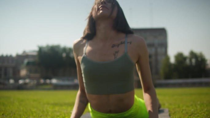 年轻女子穿着霓虹绿色运动短裤在绿草地上做瑜伽伸展运动。体育场上的女运动员射击。户外静坐冥想