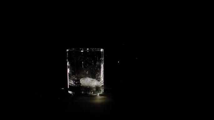冰块正落入黑色背景的玻璃杯中。三块碎冰掉进空杯子里。