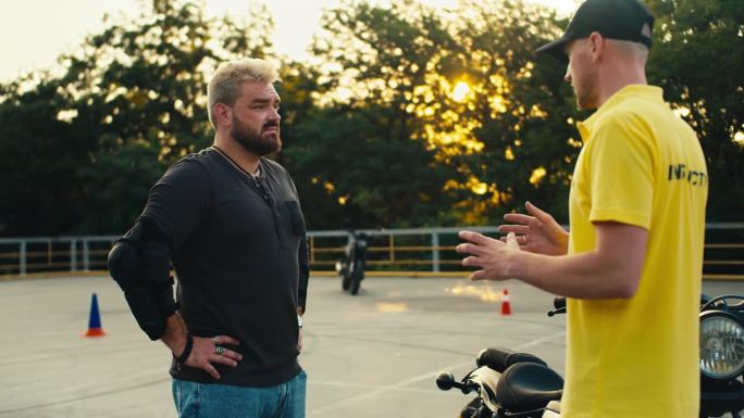一名身穿灰色运动衫、蓄着浓密胡须的男子专心地听着一名身穿黄色t恤的驾驶教练讲课。驾驶摩托车前的指导