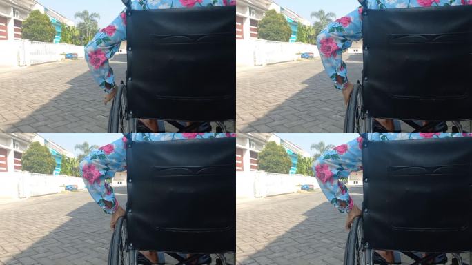 特写残疾妇女握住轮椅轮子的手。后视图残疾老年妇女坐在轮椅上晒太阳，早晨在屋前的街道上铺路