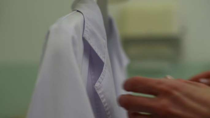 特写镜头，无法辨认的女医生从衣架上脱下白大褂，自己穿上。专业从业者准备工作