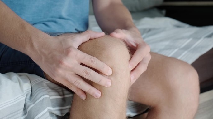 膝盖疼痛的男人坐在沙发上。男性按摩疼痛的膝盖，伤害从半月板疼痛，关节扭伤或关节炎
