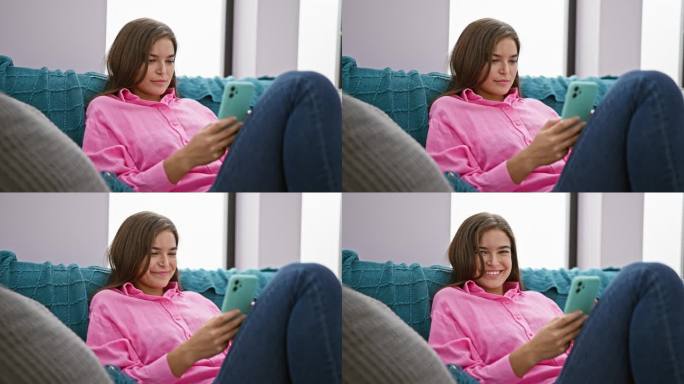 快乐的年轻西班牙女子，散发着幸福的光芒，坐在客厅的沙发上，在手机上打出欢快的信息，在家享受在线互动