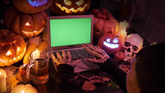 一名男子戴着骷髅手套，用彩色键在笔记本电脑键盘上打字，周围是万圣节雕刻的南瓜装饰