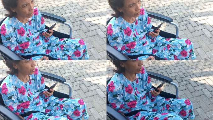 老年残疾妇女坐在轮椅上一边玩手机一边晒日光浴，早晨在屋前铺路