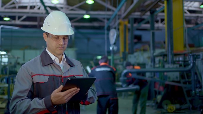 工程师在重工业工厂建筑工厂使用平板电脑在后台工作。安全帽工业检测生产制造专业男性员工
