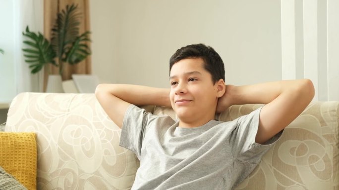 一个穿着灰色t恤又累又伤心的少年坐在沙发上看电视。无聊的男孩把手放在头上。