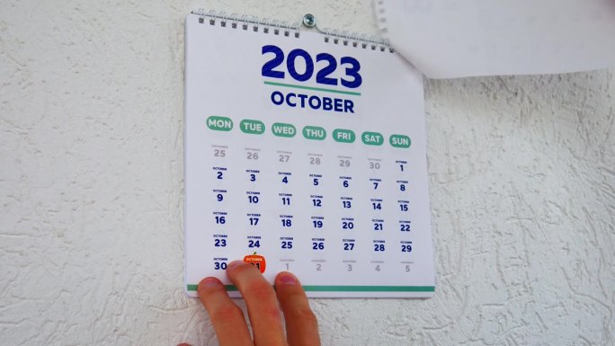 特写:两只男性的手撕掉了墙上的日历，2023年9月的那一页，紧接着是下一页，上面写着万圣节的日期
